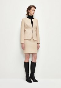Giacca da abito beige con un design aderente, abbinata a una gonna coordinata. Indossata con un top nero a collo alto e stivali neri al ginocchio. Texture liscia.