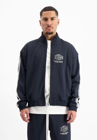 Navy zip-up jack in een gladde stof met witte strepen op de mouwen, met een logo op de linkerborst en een relaxte pasvorm.