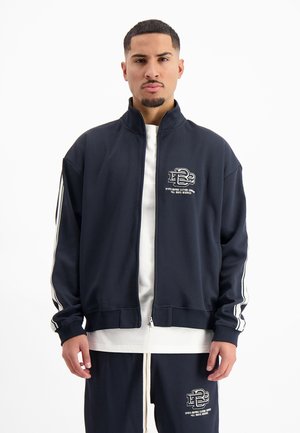 Marineblauer Zip-Jacke aus glattem Stoff mit weißen Streifen an den Ärmeln, mit einem Logo auf der linken Brust und einem lässigen Schnitt.