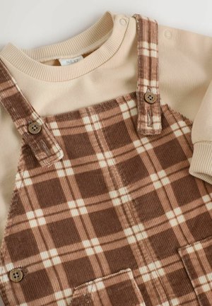 Salopette en corduroy marron avec un haut à manches longues de couleur crème, présentant un motif à carreaux, des boutons et un col en ribbed doux.