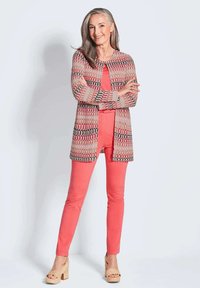 Strickjacke mit mehrfarbigem Wellenmuster, kombiniert mit einem korallenfarbenen Top und einer Hose. Plattform-Sandalen mit geflochtenen Akzenten. Weiche Texturen.