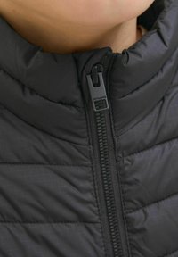 Jachetă neagră căptușită cu guler înalt, având un fermoar frontal și o textură netedă. Cusăturile brodate adaugă detalii țesăturii.