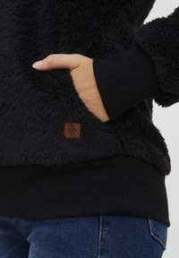 Black fleece pullover with a textured, fluffy exterior; includes a front pocket, ribbed cuffs, and a small brown brand tag.