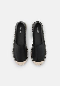 Pár černých espadrilek na slip-on s pletenými béžovými špičkami a šitými okraji, název značky "Pieces" viditelný uvnitř každé boty.