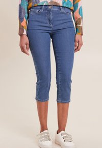 Pantalons capri en denim bleu clair. Caractéristiques d'un design ajusté, d'une coupe au niveau des genoux et de coutures contrastées. Associés avec des baskets blanches.
