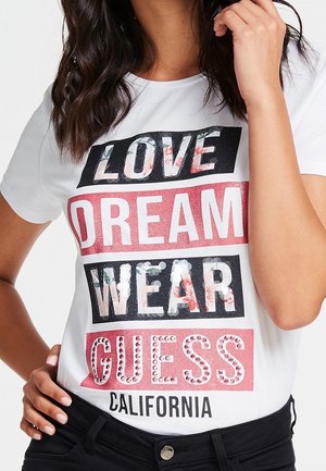Kobieta nosząca białą koszulkę z kwiatowym i brokatowym napisem ""LOVE DREAM WEAR GUESS CALIFORNIA"" na czarnych i różowych panelach.