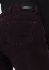 Kordhosen in tiefem Lila mit strukturierter Oberfläche. Besitzt ein schwarzes Leder-Logo und eine einzelne rückwärtige Tasche in abgerundeter Form.