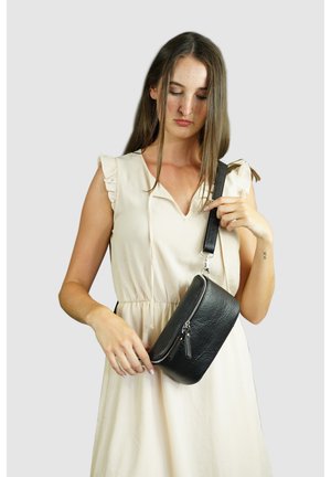 Zwarte leren crossbody tas met een gestructureerd oppervlak, ritssluiting en afneembare schouderband, vastgehouden door een persoon in een lichte jurk.
