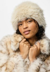 Cappello e cappotto in pelliccia sintetica di un leggero beige con un tessuto morbido e textured. Il cappotto presenta un motivo maculato. La modella tiene il colletto con le mani.