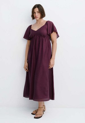 Mujer con el pelo corto y castaño posa frente a una pared blanca, lleva un vestido suelto de color morado hasta los tobillos y sandalias marrones con tira al tobillo.