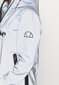 Jasnoniebieska, wodoodporna kurtka zapinana na zamek z kapturem, z logo z przodu, kieszeniami na zamki po bokach i emblematem na lewym ramieniu.