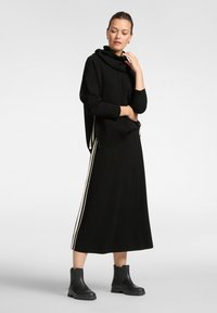 Zwarte sweatshirt met een hoge kraag en zijsplitsen, gecombineerd met een bijpassende zwarte rok met witte zijstrepen, gedragen met zwarte enkellaarsjes.