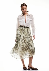 Falda fluida y plisada con un patrón abstracto en verde claro y blanco; combinada con una camisa blanca transparente de botones y zapatos planos de malla oscuros.