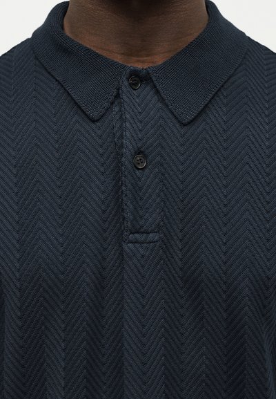 Κοντινό πλάνο σε σκούρο μπλε ριγωτό πλεκτό πόλο πουκάμισο με μυτερό γιακά και δύο μαύρα κουμπιά, φορεμένο από άτομο με σκούρο δέρμα.