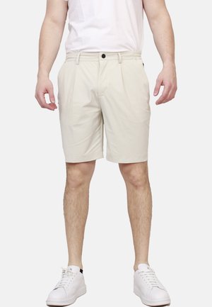 Uomo che indossa pantaloncini beige fino al ginocchio, scarpe da ginnastica bianche e una maglietta bianca in piedi davanti a uno sfondo semplice.