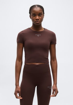 Edzett nő, aki barna rövid ujjú crop topot és hozzáillő, magasderekú leggingset visel, mindkettőn "HYROX" és Puma logók szerepelnek, haját fonva viseli.