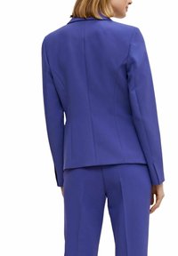 Skräddarsydd blå blazer med kavajslag, markerade axlar och en figursydd silhuett. Mjukt tyg och elegant sömnadsdetaljering.