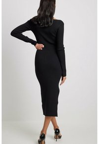 Svart ribbad stickad midiklänning med långa ärmar och figurnära siluett, med en rundad rygg och slät yta. Svarta högklackade skor.