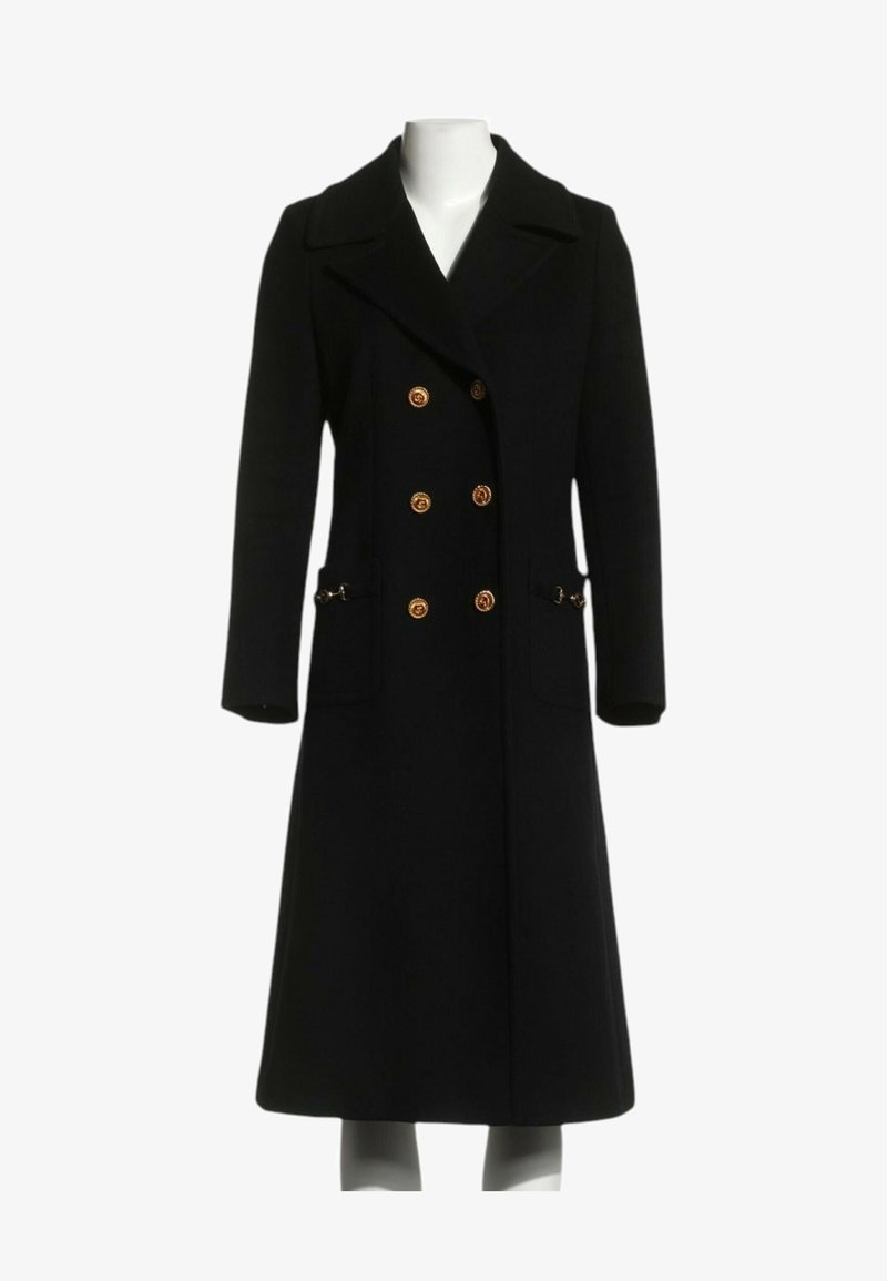 Long manteau noir croisé avec boutons dorés, larges revers, poches avant et manches longues sur un mannequin sans tête.
