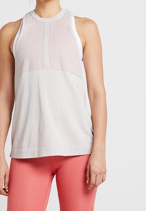 Kvinde iført lysegrå ærmeløs netagtig træningstop og koralrosa leggings, stående mod en ensfarvet grå baggrund.