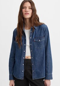 Chemise en denim bleu foncé, avec un devant à boutons, deux poches poitrine et un col classique. Manches longues avec des poignets boutonnés.