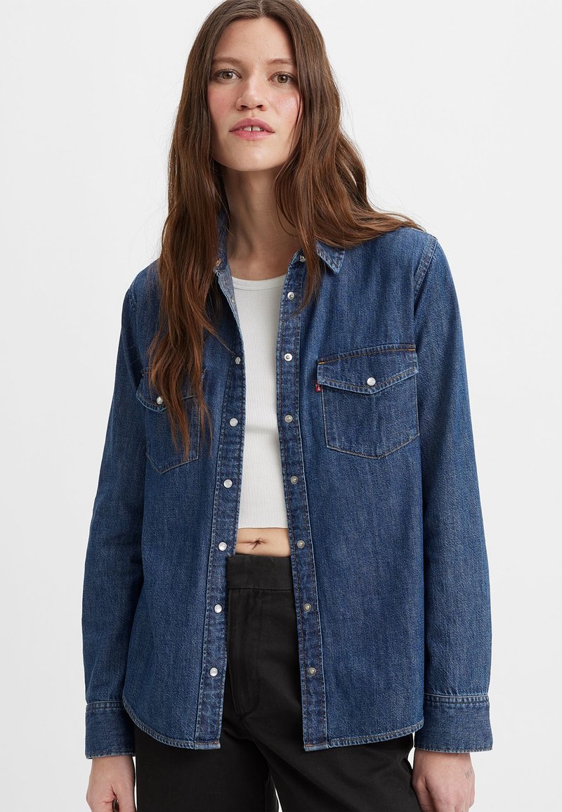 Chemise en denim bleu foncé, avec un devant à boutons, deux poches poitrine et un col classique. Manches longues avec des poignets boutonnés.