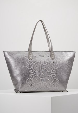 Srebrna torba typu tote z wytłaczanym kwiatowym i ornamentowym wzorem, dwoma rączkami oraz małymi metalowymi ćwiekami, na białej powierzchni.
