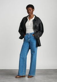 Schwarze Leder-Übergrößejacke, weißes gestricktes Polohemd mit horizontalen Streifen, blaue weit geschnittene Jeans und braune flache Schuhe, stehend auf einem grauen Boden.