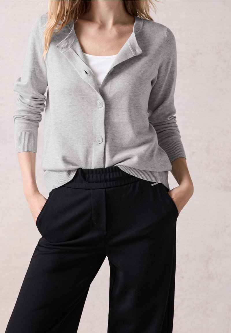 Femme portant un pull gris clair boutonné sur un haut blanc avec un pantalon noir à taille élastique, mains dans les poches, debout devant un fond neutre.