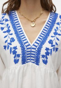 Vit blus med intrikata blå blommönsterbroderier och en texturerad V-ringning. Tyget är lätt och har en rynkad fåll.