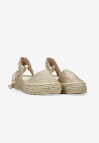 Espadrilles beige trenzadas con suela acolchada, que presentan lazos en el tobillo y una superficie texturada. Material natural y diseño redondeado.