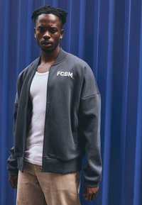 Graue Bomberjacke mit Reißverschluss, gerippten Bündchen und dem auf der Brust gestickten Text "FCBM", getragen über einem weißen Tanktop.