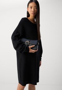 Black leather handbag with a structured design, featuring a flap closure and a gold-tone buckle detail, held close to a model in a black dress.