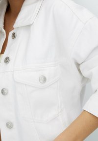 Bílá denimová bunda s kovovými patentkami, náprsní kapsou s klopou a ohrnutými rukávy, zdůrazňující texturovaný design látky.