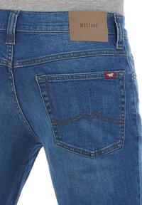 Jeans de mezclilla en azul oscuro con una textura suave. Incluye un parche de cuero etiquetado "MUSTANG" y una marca roja en el bolsillo trasero.