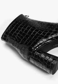 Zapato de tacón alto de charol negro con patrón de cocodrilo, que presenta un diseño curvado y un tacón grueso. Líneas limpias y acabado brillante.