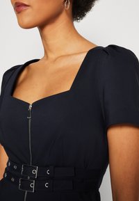 Femme portant une robe noire avec un col carré, des manches courtes bouffantes, une fermeture éclair à l'avant et une ceinture à double boucle, montrée des épaules à la taille.