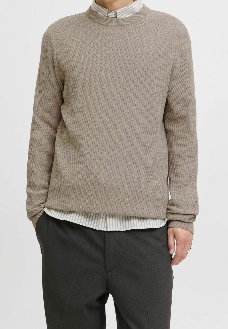 Personne portant un pull beige texturé par-dessus une chemise blanche rayée et un pantalon foncé, debout les mains partiellement dans les poches.