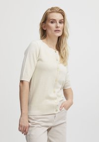 Lys beige cardigan lavet af blødt stof, med korte ærmer, rund udskæring og guldknapper foran. Glat tekstur.