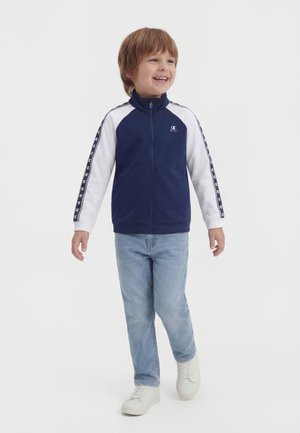 Χαμογελαστός νεαρός αγόρι περπατά, φοράει ναυτικό μπλε και λευκό τζάκετ με φερμουάρ, με λογότυπο στα μανίκια και το στήθος, ανοιχτό μπλε τζιν και λευκά αθλητικά παπούτσια.
