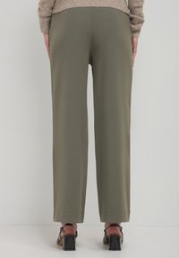 Pantaloni a gamba larga verde oliva, texture liscia, con orlo pulito, abbinati a scarpe con tacco quadrato nere e lucide. Maglione tricot marrone chiaro visibile.
