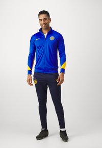 Giacca sportiva blu con dettagli gialli e logo, abbinata a pantaloni blu navy. Presenta una chiusura frontale con zip e polsini aderenti, indossata con scarpette nere.