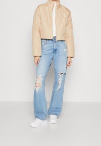 Beżowa pikowana krótka kurtka z wysokim kołnierzem, noszona na białym swetrze. Jasnoniebieskie, szerokie dżinsy z przetarciami, z dziurami na kolanach. Białe trampki.