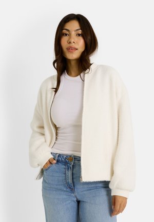 Cárdigan blanco, texturizado, de apertura delantera con hombros caídos y puños acanalados, usado sobre una blusa ajustada gris claro y jeans de tiro alto azules.