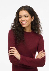 Top a maniche lunghe color marrone scuro con collo tondo a costine. Tessuto leggero con silhouette aderente. Capelli ricci visibili, braccia incrociate davanti.