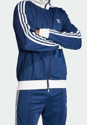 Personne portant un survêtement Adidas bleu avec des bandes blanches sur les manches et le pantalon, bras croisés, avec une fermeture éclair à l'avant de la veste.