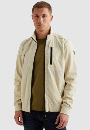 Jeune homme portant une veste beige zippée sur une chemise vert olive, debout devant un fond clair uni.