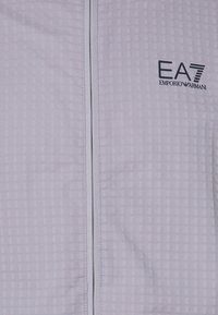 Svijetlo siva jakna sa zatvaračem napravljena od teksturirane tkanine, s suptilnim uzorkom mreže i crnim EA7 Emporio Armani logom na grudima.