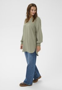 Bluza verde măsliniu cu mâneci lungi, din țesătură texturată, cu decolteu inelar și manșete elastice, asortată cu blugi evazați albaștri și pantofi maro.