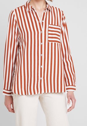 Femme portant une chemise boutonnée à manches longues avec des rayures verticales rouille et blanches et une poche poitrine à rayures horizontales.